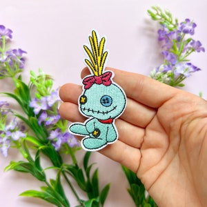 Peut inclure: Un patch brodé bleu et jaune à repasser de Stitch de Lilo & Stitch. Stitch est assis, les bras croisés, et porte un nœud rose sur la tête.