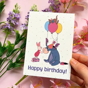 May include: A greeting card with a cartoon illustration of Winnie the Pooh characters. Piglet offers a gift to Eeyore, who is wearing a party hat and holding balloons with Roo and Kanga. The words "Happy birthday!" are printed on the card.