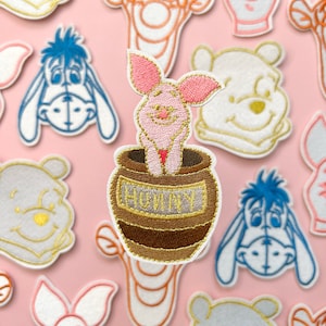 May include: Embroidered iron-on patch featuring a pink piglet character from Winnie the Pooh, standing in a brown honey pot with the word "HUNNY" in gold lettering. The patch is surrounded by other embroidered patches of Winnie the Pooh characters.