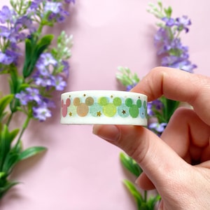 Op de afbeelding: Een rol decoratieve washi tape met een herhalend patroon van Mickey Mouse silhouetten in pastelkleuren, waaronder roze, oranje, groen en blauw, met gouden ster accenten. De tape wordt tegen een roze achtergrond met paarse bloemen gehouden.
