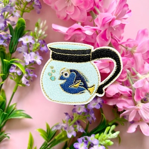 May include: A blue and yellow embroidered patch of Dory the fish from Finding Nemo, inside a blue and white coffee pot.