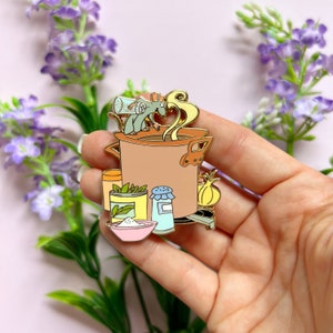 Puede incluir: Un pin de esmalte dorado con un ratón de dibujos animados revolviendo una olla de sopa con varios ingredientes, incluyendo un frasco de especias, un salero, un tazón de harina y una cebolla.