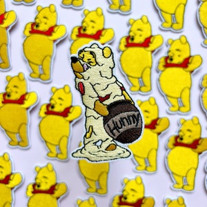 Pode incluir: Um remendo bordado do Winnie the Pooh com o Pooh coberto de mel e segurando um pote de mel que diz "Hunny". O remendo é cercado por outros remendos do Winnie the Pooh.
