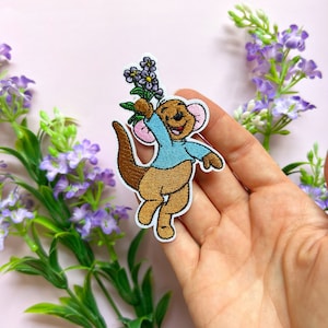 May include: An embroidered iron-on patch featuring a cartoon kangaroo holding a bouquet of purple flowers. The kangaroo is brown with a blue shirt and pink ears.