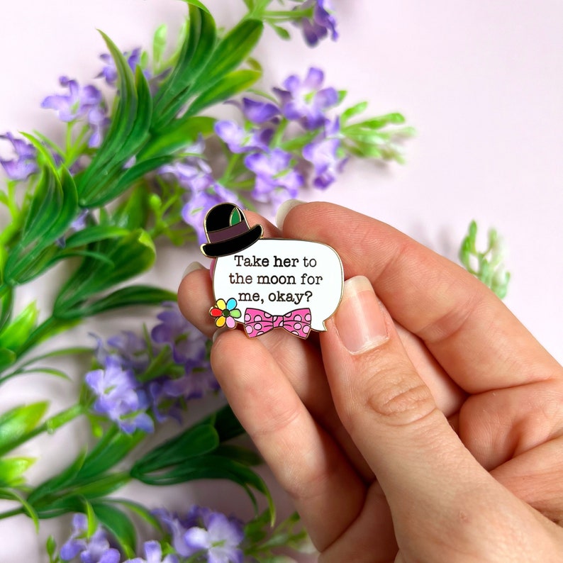 May include: A white enamel pin in the shape of a speech bubble with the text "Take her to the moon for me, okay?" A black top hat, a pink bow tie, and a colorful flower are also featured on the pin. The pin is held in a hand.