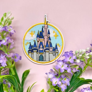 Op de afbeelding: Ronde geborduurde patch met een sprookjeskasteel met blauwe torens, roze details en een gouden torenspits, tegen een lichtblauwe lucht met gele sterren. Perfect om toe te voegen aan kleding of accessoires.