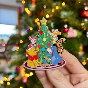 Puede incluir: Un colorido pin de esmalte con Winnie the Pooh y sus amigos reunidos alrededor de un árbol de Navidad. El pin incluye a Piglet, Pooh, Tigger, Eeyore y una pequeña figura. El árbol está decorado con adornos y coronado con una estrella dorada.