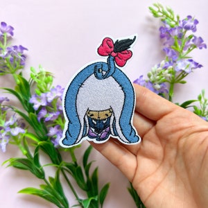 May include: Embroidered iron-on patch featuring a blue, white, and pink cartoon character with a bow. The character is hiding under a blue blanket.