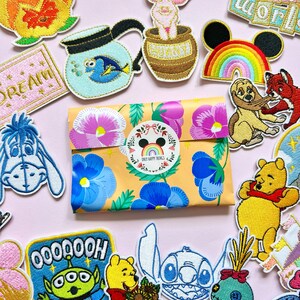 Puede incluir: Una colección de parches bordados con personajes de dibujos animados y diseños florales. En el centro hay un paquete envuelto con un estampado floral y una pegatina circular. Los parches incluyen personajes de Disney y un arcoíris.