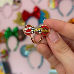 May include: A metal enamel pin shaped like mouse ears featuring a red race car face on the left ear, a Rust-eze logo on the right ear, and a black and white checkered bow in the center with the number 95 and a yellow lightning bolt design.