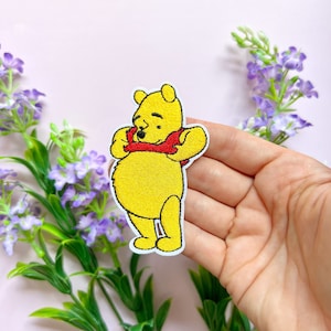 Op de afbeelding: Gestrikte strijk-op patch met Winnie de Poeh die een rode reddingsvest draagt. De patch is geel en wit met zwarte gezichtskenmerken.