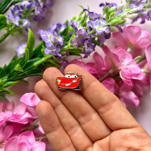 Op de afbeelding: Een rode emaille pin met een cartoonauto met een lachend gezicht. De pin heeft zwarte banden en het nummer 95 aan de zijkant. De pin staat tegen een achtergrond van roze en paarse bloemen.