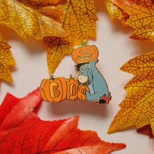 Puede incluir: Un pin temático de Halloween con Eeyore de Winnie the Pooh, rodeado de calabazas naranjas y hojas de otoño. Las calabazas deletrean "BOO". El pin está sobre un fondo de colorido follaje otoñal.