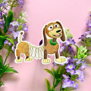 Op de afbeelding: Geborduurde strijkpatch van Slinky Dog uit de Toy Story-franchise. De patch toont Slinky Dog in zijn kenmerkende bruin en tan kleuren met een gouden veerlichaam en een groene halsband.