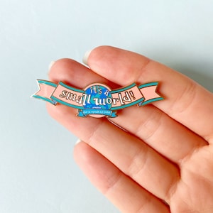 Puede incluir: Un pin de esmalte rosa y azul con borde dorado. El pin presenta un globo terráqueo con el texto "it's a small world!" y la frase francesa "que le monde est petit" en una pancarta.