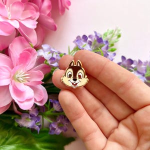 May include: A Chip enamel pin depicting a smiling Chipmunk face in brown, tan, and white, with a gold-coloured outline. The pin is held in a hand, with pink and purple flowers in the background.