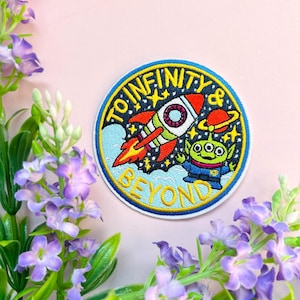 Op de afbeelding: Een ronde geborduurde patch met een blauwe, gele en rode raket en een groene alien. De tekst "To Infinity & Beyond" is in geel rond de rand van de patch geschreven.