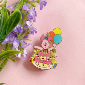 Op de afbeelding: Een gouden emaille pin met een roze varken dat een feestmuts draagt en ballonnen vasthoudt. Het varken zit op een roze en gele verjaardagstaart met kaarsen en cadeautjes.