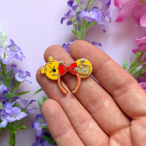 Op de afbeelding: Een goudkleurige emaille speld in de vorm van een haarband met oren. Eén oor toont een geel berengezicht, het andere een honingpot met het label HONEY, verbonden door een rode strik. Het ontwerp bevat kleine bijtjes en oranje accenten op de band.