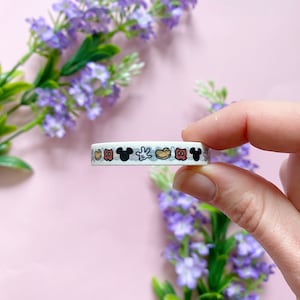 Op de afbeelding: Een rol witte washi tape met een herhalend patroon van Mickey Mouses hoofd, handschoenen, schoenen en shorts in zwart, rood en geel. De tape wordt door een hand vastgehouden tegen een roze achtergrond met paarse bloemen.