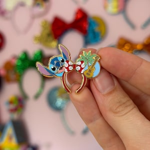 Puede incluir: Un pin de esmalte metálico con forma de orejas que presenta un personaje alienígena azul a la izquierda, un lazo con estampado floral rojo y blanco en el centro, y una palmera verde con un ukelele marrón a la derecha sobre un fondo azul claro.