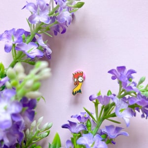 Op de afbeelding: Een kleurrijke emaille pin met een cartoon haan met een rode bril, een gele body en een roze kop. De pin is omgeven door paarse bloemen en groene bladeren tegen een lichtpaarse achtergrond.