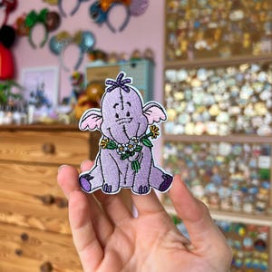 Könnte beinhalten: Ein lavendelfarbener, gestickter Aufnäher mit einer Cartoon-Elefantenfigur mit einem Blumengirlande. Der Aufnäher hat einen lila Umriss und wird in einer Hand gehalten. Der Hintergrund umfasst Disney-Themen-Haarbänder und Anstecknadeln.