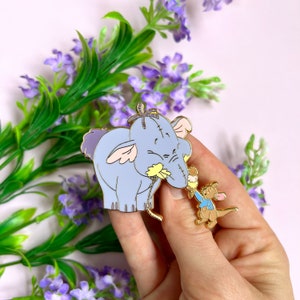 Pode incluir: Um conjunto de pinos de esmalte dourado com personagens do Winnie the Pooh. O pino maior mostra um elefante azul com uma expressão triste, enquanto o pino menor mostra um canguru amarelo segurando um balão azul.