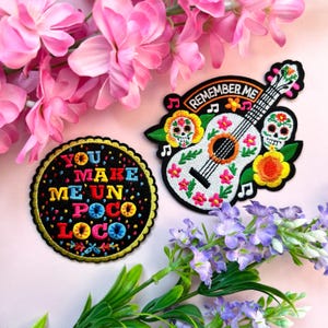 May include: Two embroidered iron-on patches. One patch is round and features the text "You Make Me Un Poco Loco" in colorful letters on a black background. The other patch is shaped like a guitar and features a white guitar with colorful flowers and two sugar skulls. The text "Remember Me" is above the guitar.