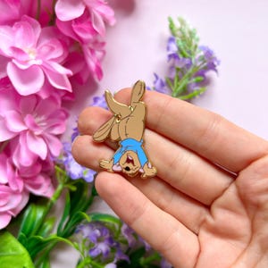 Peut inclure: Une épingle en émail dorée représentant un personnage de dessin animé en équilibre sur les mains. Le personnage a une fourrure beige, une chemise bleue et sourit. L'épingle est tenue dans une main, avec des fleurs roses et violettes en arrière-plan.