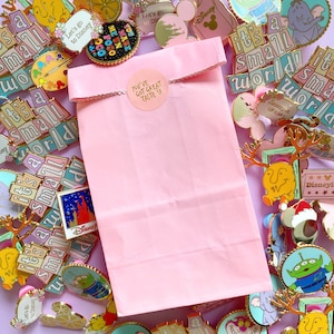 Op de afbeelding: Een roze papieren tas met een gouden sticker waarop "You've got great taste" staat, omringd door een verscheidenheid aan kleurrijke Disney-thema enamel pins. De pins tonen personages en zinnen uit populaire Disney-films en -attracties, waaronder Mickey Mouse, Winnie de Poeh en "It's a Small World."