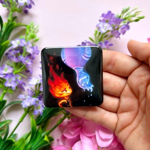 May include: A square, black pin depicting two cartoon characters: a red, fiery character and a blue, watery character. The figures are set against a dark background. The pin is held in a hand, with purple flowers and pink petals in the background.