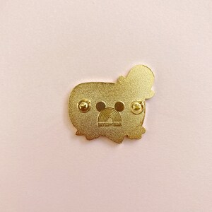 May include: A gold-colored enamel pin featuring a cartoon character with a hat. The pin has two pin backs and a rainbow design with the text "DAILYHAPPYTHINGS". The pin is on a pink background.