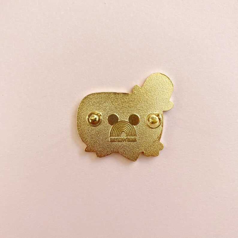 May include: A gold-colored enamel pin featuring a cartoon character with a hat. The pin has two pin backs and a rainbow design with the text "DAILYHAPPYTHINGS". The pin is on a pink background.