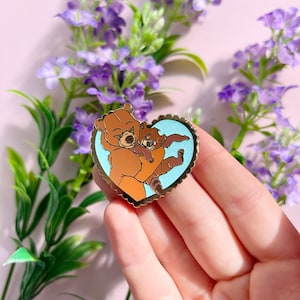 Peut inclure: Épingle en émail en forme de cœur représentant deux ours bruns enlacés. L'épingle a une bordure dorée avec un bord festonné et un fond bleu clair. Les ours sont de style dessin animé.