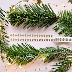 Pode incluir: Dois rolos de fita washi com um design de enfeites de Natal marrons e dourados. A fita está sobre um fundo rosa com ramos de pinheiro verdes e luzes de corda douradas.