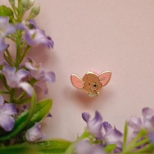 Peut inclure: Une petite broche en émail métallique représentant la tête d'un personnage de kangourou de dessin animé avec de grandes oreilles roses, un visage beige et un petit collier bleu, centrée sur un fond rose uni avec des fleurs violettes.