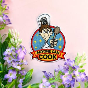 Può includere: Toppa ricamata da stirare con un disegno di un cuoco dei cartoni animati che tiene un cucchiaio e il testo "Anyone Can Cook".