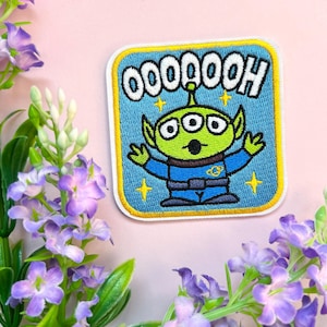 Op de afbeelding: Een blauwe en groene geborduurde patch met een drie-ogig alienfiguur met de tekst "OOOAAOOOH" erboven. De alien draagt een blauw ruimtepak met een gele riem en is omringd door sterren.