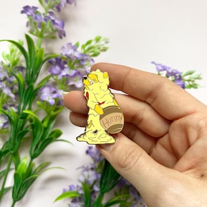 Op de afbeelding: Een gele emaille pin van een beer bedekt met honing, met een bruine honingpot. Op de honingpot staat het woord "Hunny". De pin wordt in een hand gehouden, met een achtergrond van paarse bloemen.