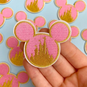 Op de afbeelding: Roze geborduurde patch in de vorm van Mickey Mouse oren, met een gouden kasteelontwerp. De patch heeft een witte rand. Verschillende vergelijkbare patches zijn zichtbaar tegen een lichtblauwe achtergrond.