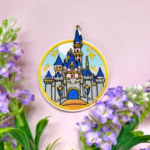 Op de afbeelding: Geborduurde patch met een sprookjeskasteel met blauwe torens, roze muren en een gouden rand. Het kasteel staat tegen een lichtblauwe lucht met gele sterren, alles binnen een gele cirkel. De patch is omgeven door paarse bloemen.
