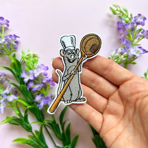 May include: Iron-on embroidered patch featuring a cartoon rat wearing a chef's hat and holding a wooden spoon. The rat is grey and the spoon is brown.