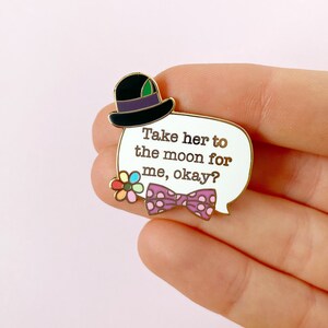 May include: A gold-edged enamel pin with a white speech bubble that reads "Take her to the moon for me, okay?" The pin features a black top hat, a purple polka-dot bow tie, and a colorful flower.