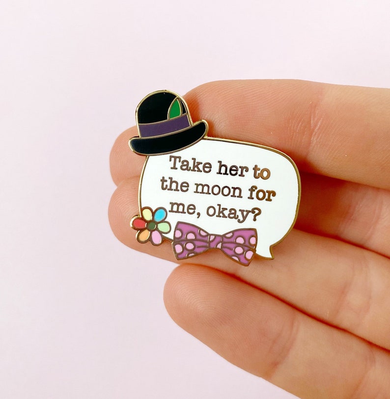 May include: A gold-edged enamel pin with a white speech bubble that reads "Take her to the moon for me, okay?" The pin features a black top hat, a purple polka-dot bow tie, and a colorful flower.