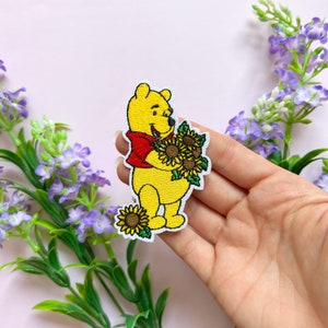 Puede incluir: Un parche bordado para planchar con Winnie de Pooh sosteniendo un ramo de girasoles. El parche es amarillo, rojo y verde.