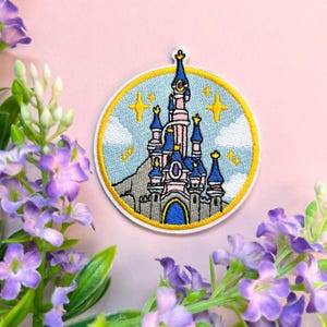 Puede incluir: Parche bordado con un castillo de cuento de hadas con torres azules, muros rosas y una puerta arqueada azul. El castillo está situado sobre un cielo azul claro con estrellas amarillas y nubes blancas, todo dentro de un borde circular dorado.