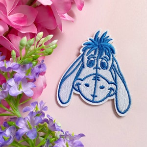 Op de afbeelding: Een geborduurde strijk-op patch in blauw en wit, met een cartoon ezel met een droevige uitdrukking. De ezel kijkt naar beneden met zijn oren die hangen.