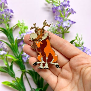 Peut inclure: Épinglette en émail doré représentant un élan de dessin animé avec un ourson sur le dos. L'élan est brun et orange avec un visage blanc et des bois. L'ourson est brun avec une poitrine blanche et s'accroche aux bois de l'élan.