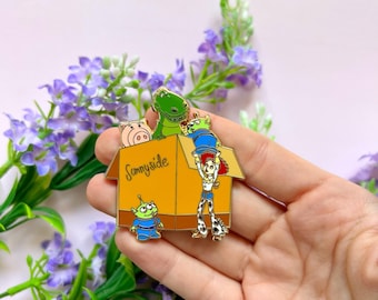 May include: A gold-colored enamel pin featuring characters from the Toy Story franchise. The characters are inside a box labeled "Sunnyside". The characters include Woody, Buzz Lightyear, Rex, Hamm, and a green alien.